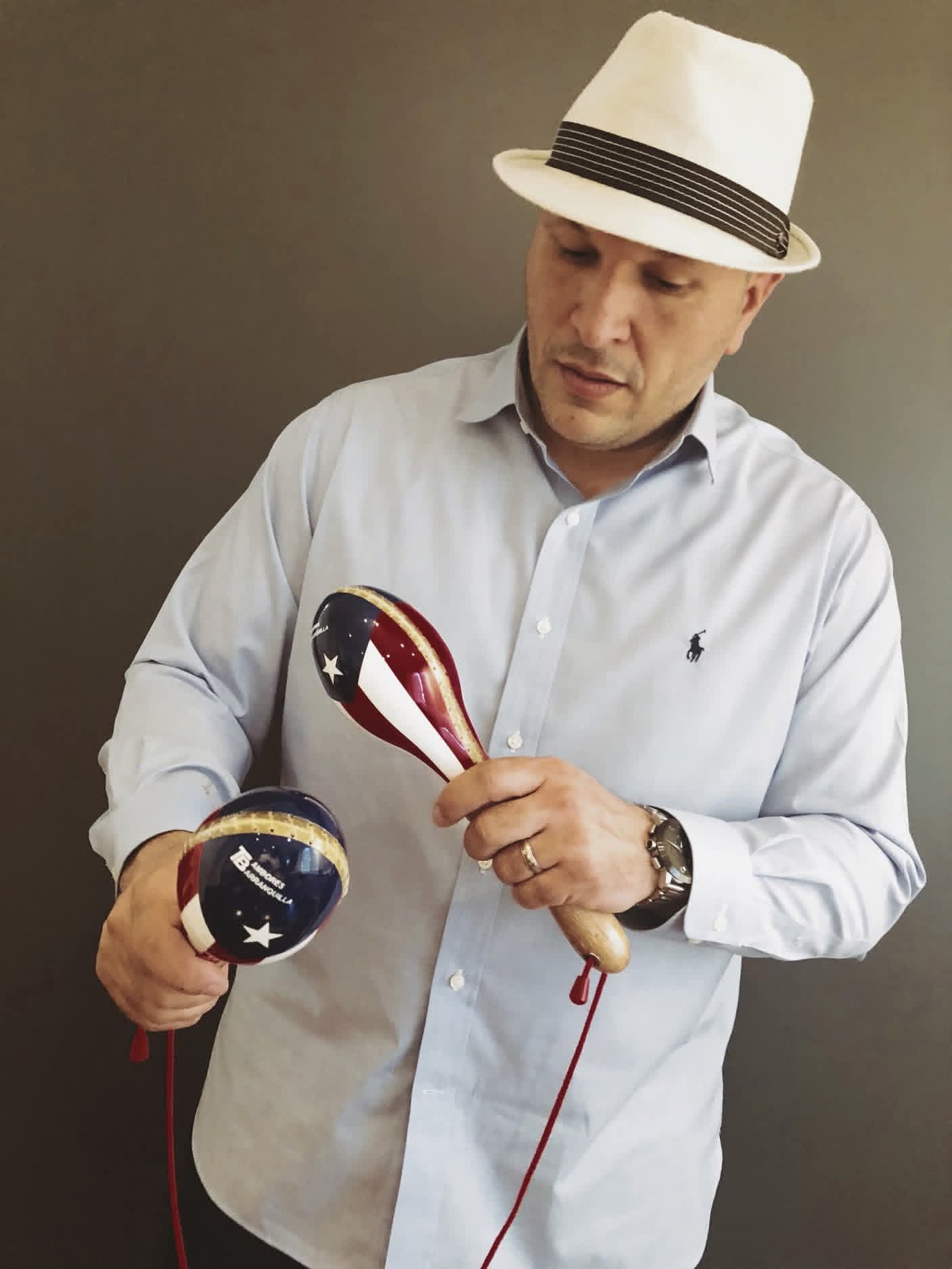 Man in a cream straw hat and pale blue shirt holding hand-painted maracas with flag designs and a Tambores Barranquilla logo against a dark wall